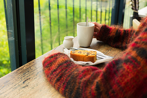 The hands of a young woman resting on a table by the window as she is having tea and cake