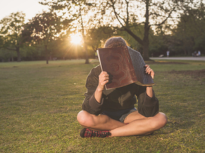 A young woman is sitting on the grass in a park at sunset and is hiding her face behind a notebook