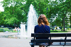 A young woman is sitting on a bench in a park and is looking at a fountain