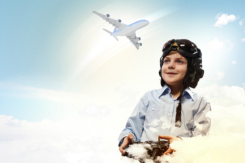 Image of little boy in pilots helmet playing with toy radiocontrolled airplane against clouds background