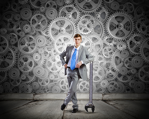 Young determined businessman leaning on wrench and cogwheels at background