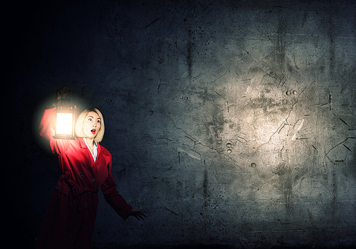 Young blonde in red cloak with lantern in darkness