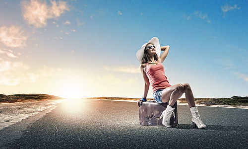 Young pretty girl traveler sitting on suitcase aside of road