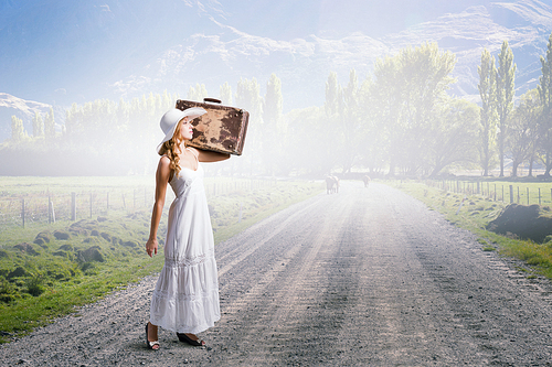 Woman with suitcase in white long dress and hat on countryside road