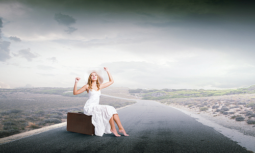 Woman in white long dress and hat sitting on her luggage on asphalt  road