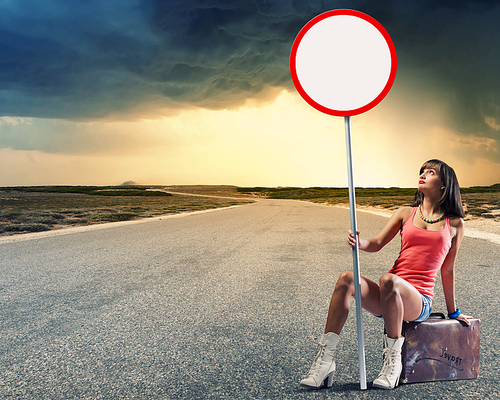 Young pretty girl traveler sitting on suitcase aside of road