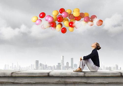 Young pretty businesswoman sitting on top of building with colorful balloons flying above