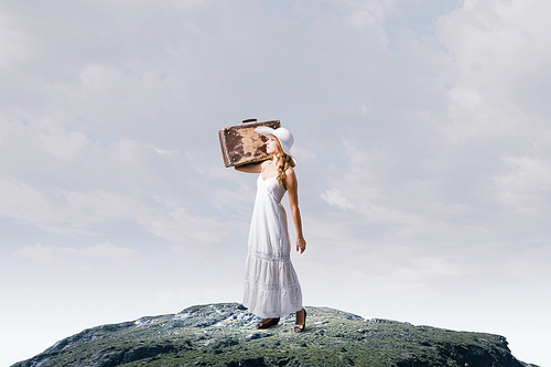 Woman with suitcase in white long dress and hat on countryside road