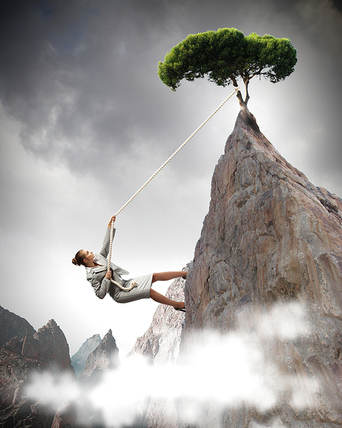 businesswoman climbing steep mountain with tree on top hanging on rope