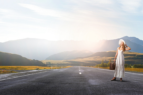 Woman with suitcase in white long dress and hat on asphalt  road