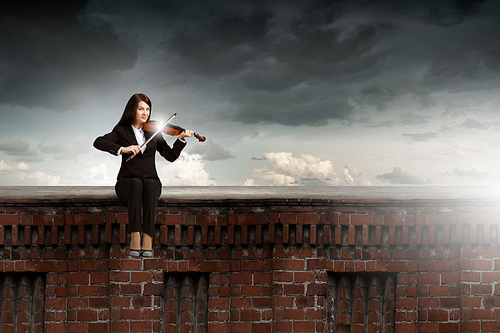 Young woman sitting on building top and playing violin
