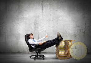 Young confident businessman sitting in chair with legs on stack of coins