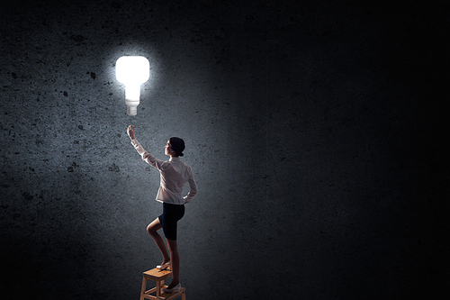 Back view of businesswoman standing on chair and reaching light bulb