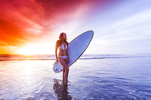 A beautiful young slim sporty woman in bikini with a surfboard is standing at ocean beach at sunset