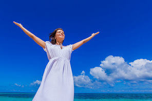 Woman in white dress posing in tropical sea beach with arms raised