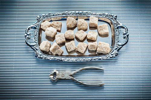 Brown sugar cubes pliers tray on striped background.