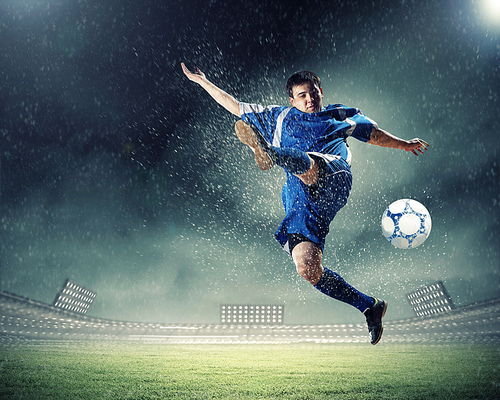 football player in blue shirt striking the ball aloft at the stadium under the rain