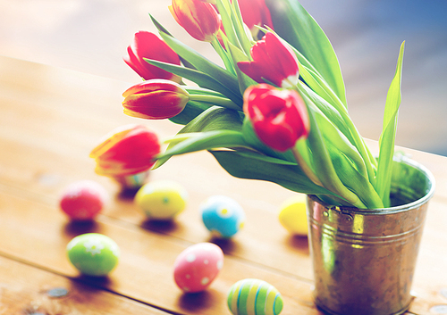 ., religious holidays and object concept - close up of tulip flowers in bucket on wooden table and colored eggs over sky background