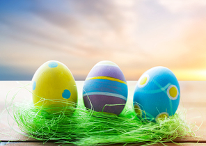 easter, religious holidays and object concept - close up of colored eggs and decorative grass on wooden surface over sky background