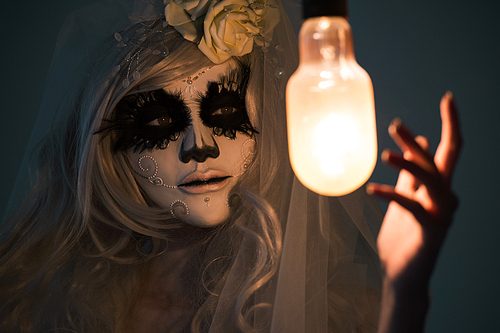 halloween witch. beautiful woman wearing 산타무에르테 mask casting spell near light bulb