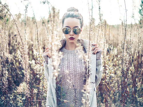 Outdoor fashion photo of young beautiful lady in autumn landscape with dry flowers. Knitted sweater, sunglusses, wine lipstick. Warm Autumn. Warm Spring