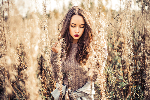 Outdoor fashion photo of young beautiful lady in autumn landscape with dry flowers. Knitted sweater, wine lipstick. Warm Autumn. Warm Spring