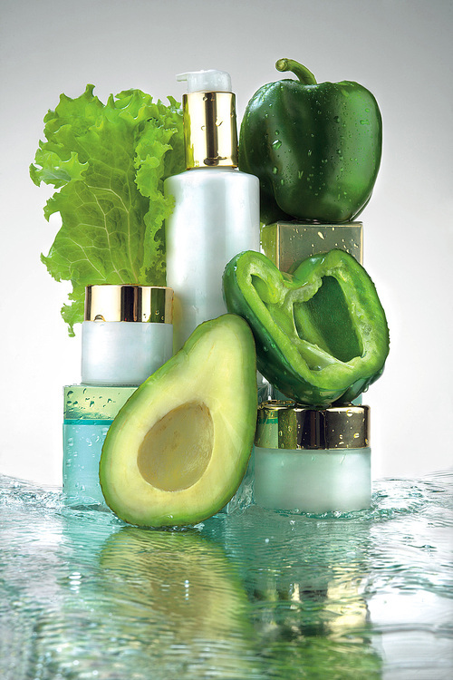 A close-up of different jars of cosmetics and green vegetables in water splash.