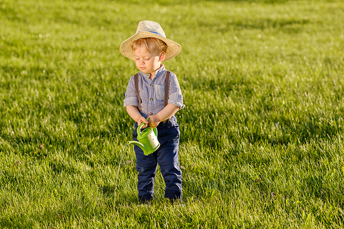 Portrait of toddler child outdoors. Rural scene with one year old baby boy wearing straw hat using watering can