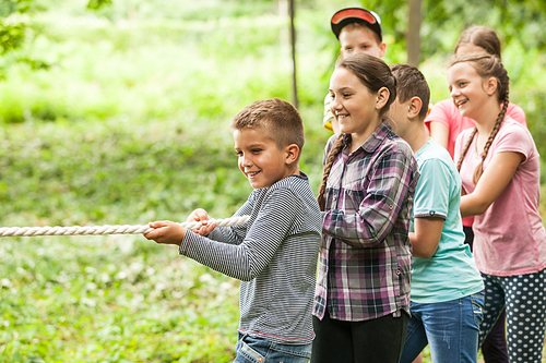 Group of happy smiling kids playing tug-of-war with rope in green park,