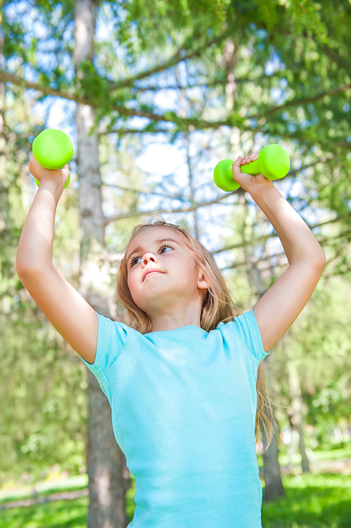 Happy little girl lifting dumbbells in park outdoors