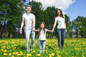 Happy family of parents and daughter walking by yellow dandelion flowers in summer park