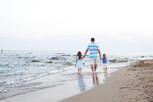 Back view of  young father and his two pretty kids walking along sand beach at evening time