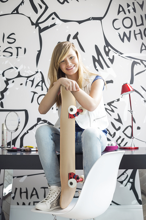 Full-length portrait of happy teenage girl with skateboard sitting on study table at home