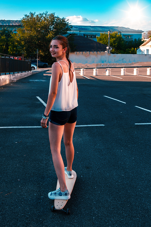 Rear view of young girl in short denim shorts with long bare legs and long hair in ponytail riding skateboard and looking back