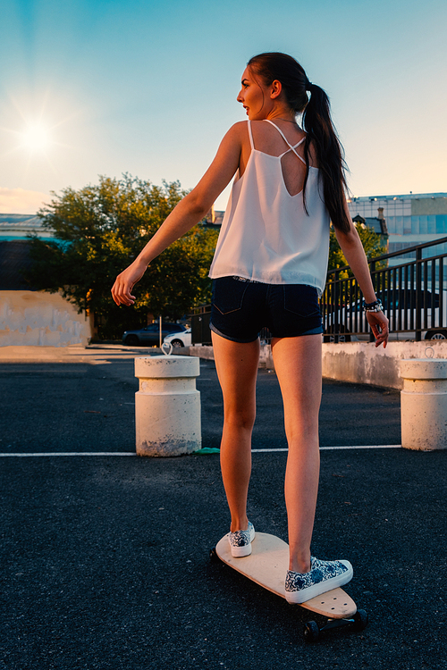 Beautiful girl riding skateboard in short denim shorts, view from back in front of early evening sky