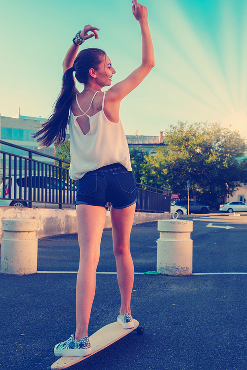 Back view of young girl in short overalls with long bare legs and long hair in ponytail riding skateboard with her hands rised, skater girl trying to keep balance