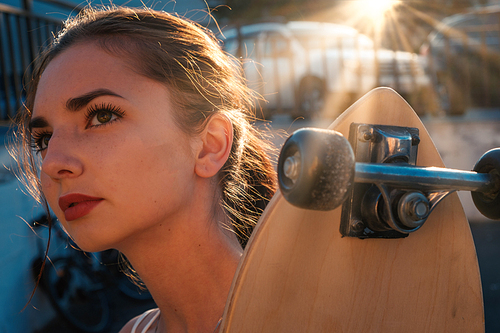 Headshot of young girl holding longboard near her face backlit by sunset light shot with sunflares
