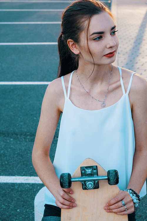 Slim girl hilding her longboard in front of her body and looking back retro color shot. On one side of model is shadow pattern and on the other - parking lot markings on background.