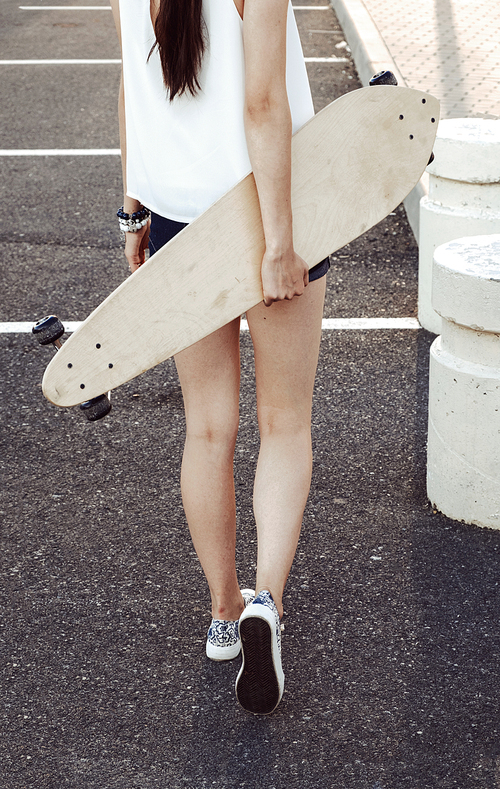 Rear view of skater girl walking away holding her longboard behind her back