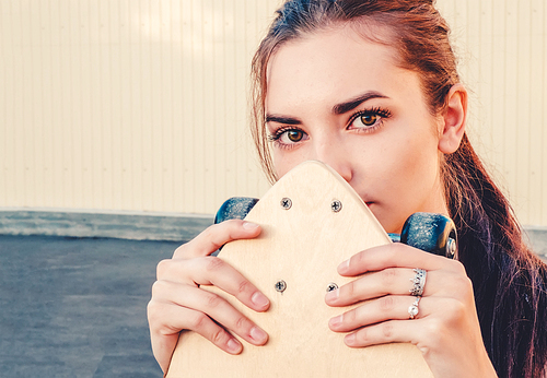 Girl holding skateboard in front of her face and . A lot of space for text on wall behind the model.