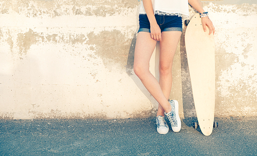 Young Girl standing near wall with her legs crossed holding her longboard leaning back weared wall, a lot of copyspace