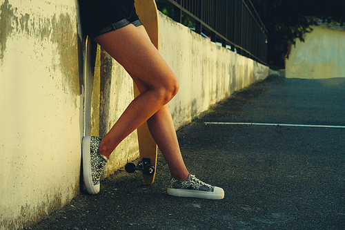 Girl with skateboard leaning back concete wall. Elegant slim legs of female longboard skater in skate park.