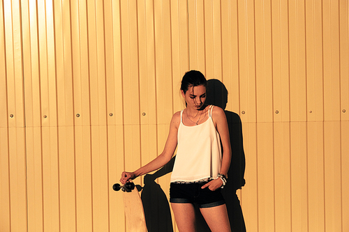 Girl in denim shorts and tank top standing with skateboard in front of yellow mwtal fence and looking down a lot of space for text