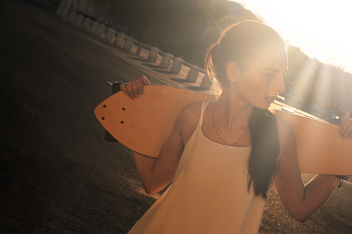 Young Woman with longboard behind her neck backlit by bright summer sun a lot of space for text