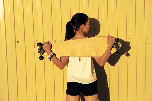 Young girl posing with her skateboard behind her back in front of yellow fence with copyspace, rear view shot