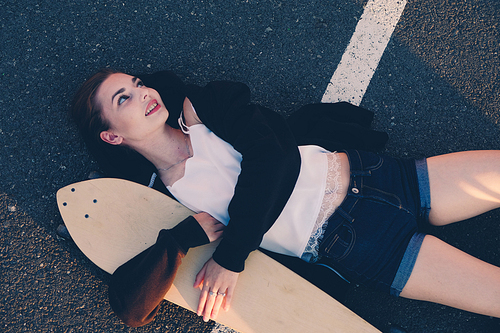 Girl lying down with her longboard on asphalt road and looking away, copyspace.