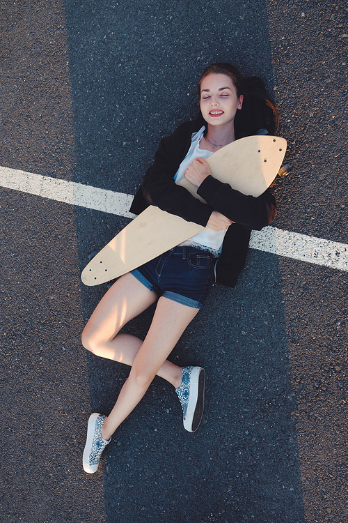 Skateg girl lying down with her longboard on asphalt street surface above view