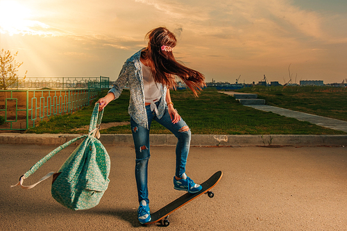 Hipster female on skateboard with backpack in her hands backlit in warm sunset light making trick on the road