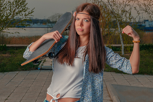 Funny girl in boho attire with skatebord on her shoulder make I-am-powerful gesture by her left hand