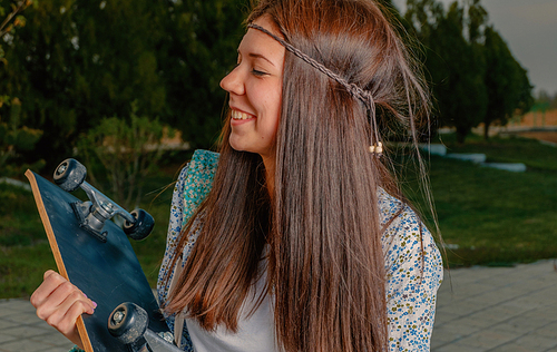 Profile View Lovely girl in boho attire with skatebord in her hands and artificial flower in her hairs looking away of camera and Smiling